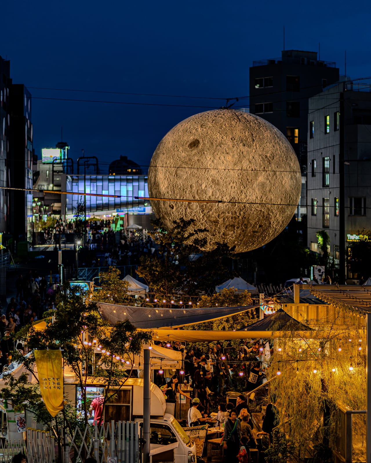 下北沢の夜空に浮かぶ大きな月のアート作品と、屋台の灯りでにぎわう下北線路街